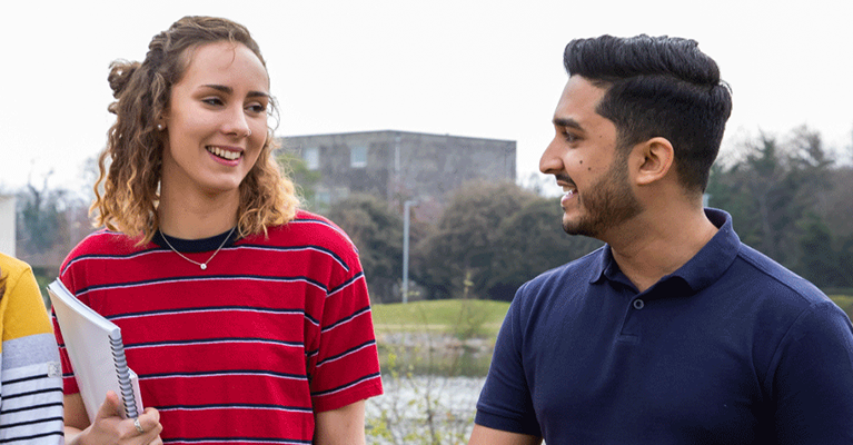 Two students walking by the upper lake on campus. The female student is holding a notebook while the male student talks to her.